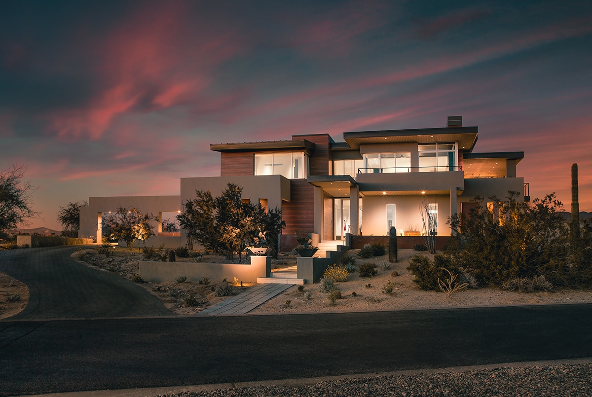 A front view of the glassy, two story home in the desert during sunset.
