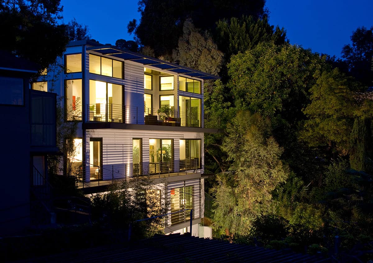 A nighttime view of the three-story home with large doors and panels of glass that lead to the tree-laden landscape.
