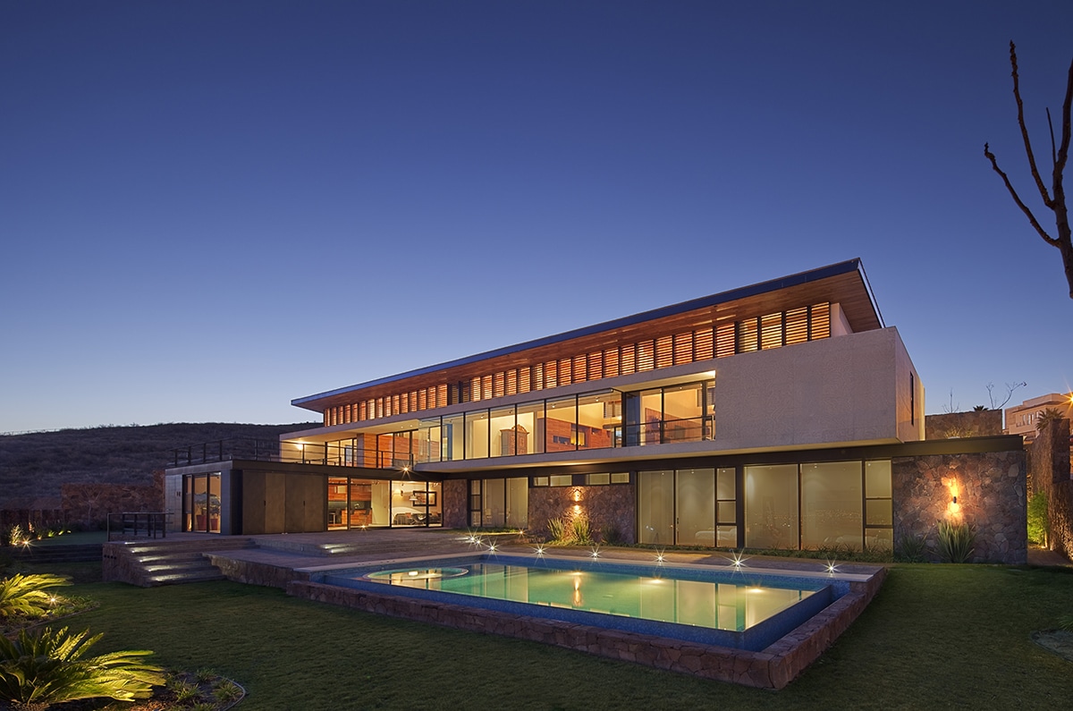 A view of the large, glassy two-story home from the backyard, showcasing lots of grass and a pool.