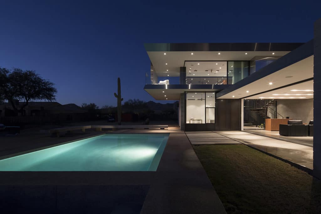 A view of the backyard with a pool at night connected to the home’s interior through windows and open walls.