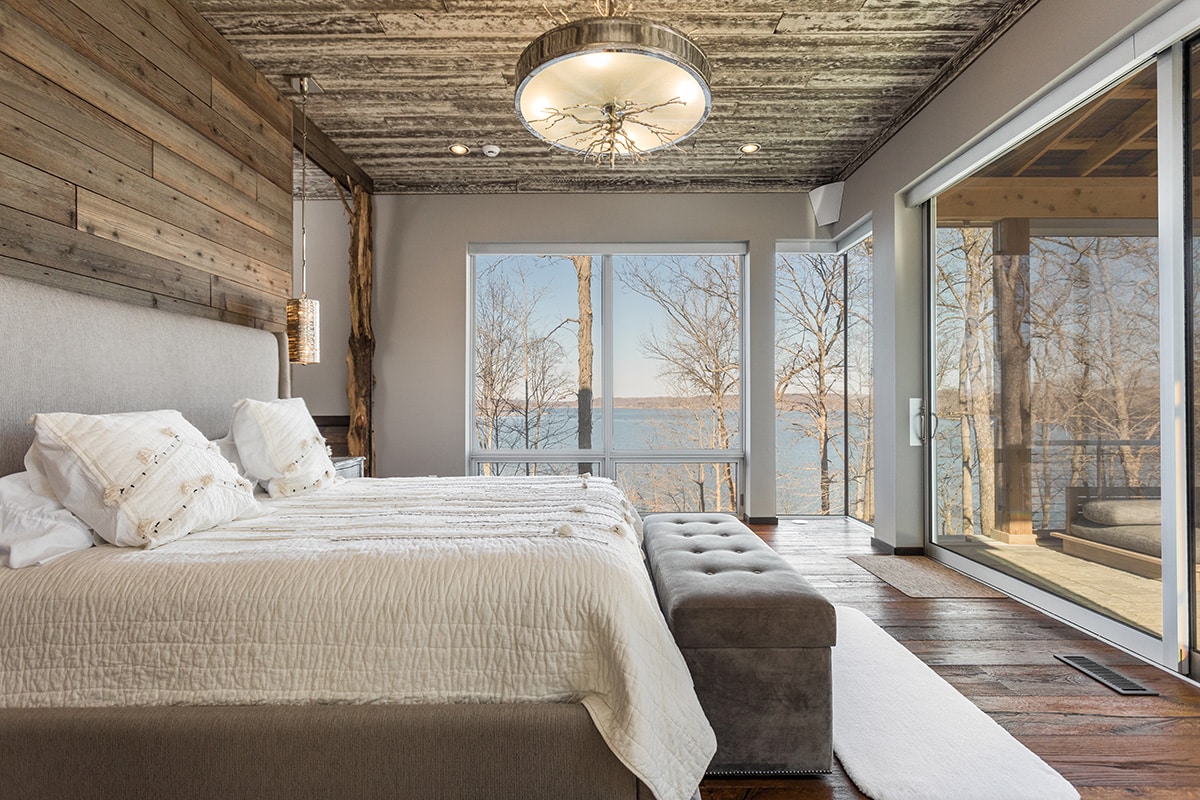 This master bedroom has a massive sliding glass door that leads to a private balcony next to windows with views of a lake.