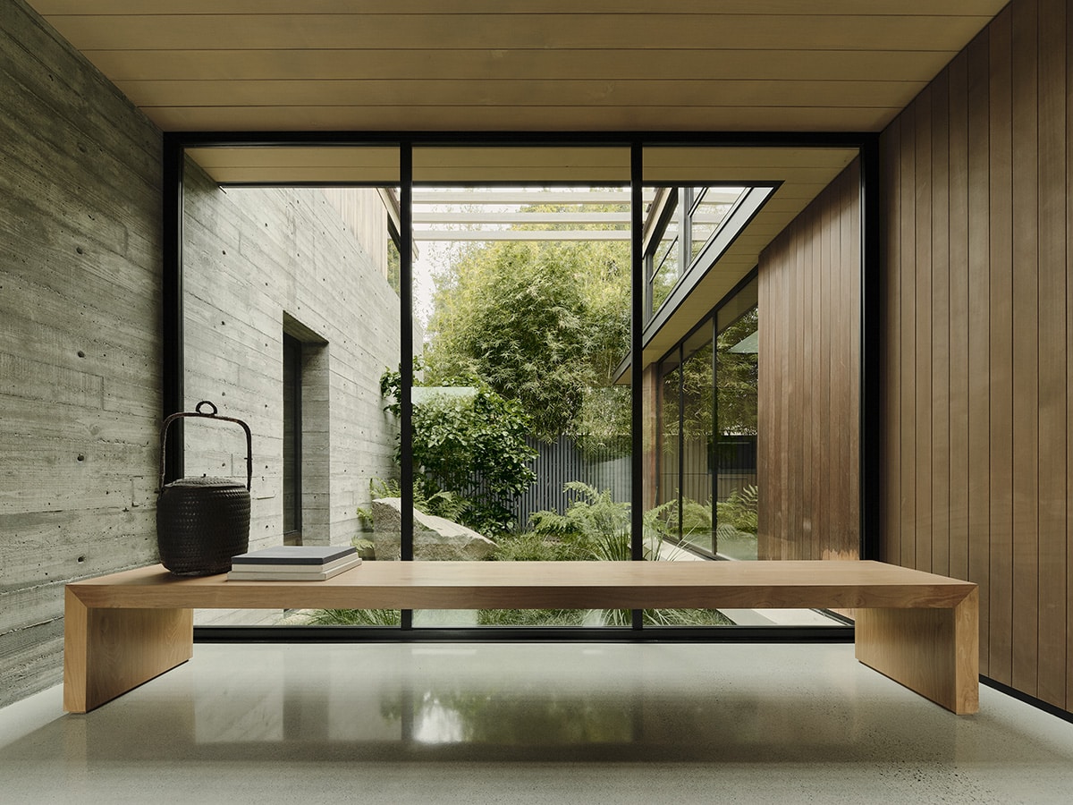 A large, wooden bench looks out into the tree-filled courtyard through wall-to-ceiling fixed windows.