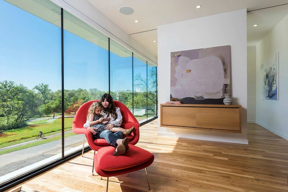 Floor-to-ceiling glass at the front of the home frames a view of the park across the street while a mother and child relax on a red chair inside.