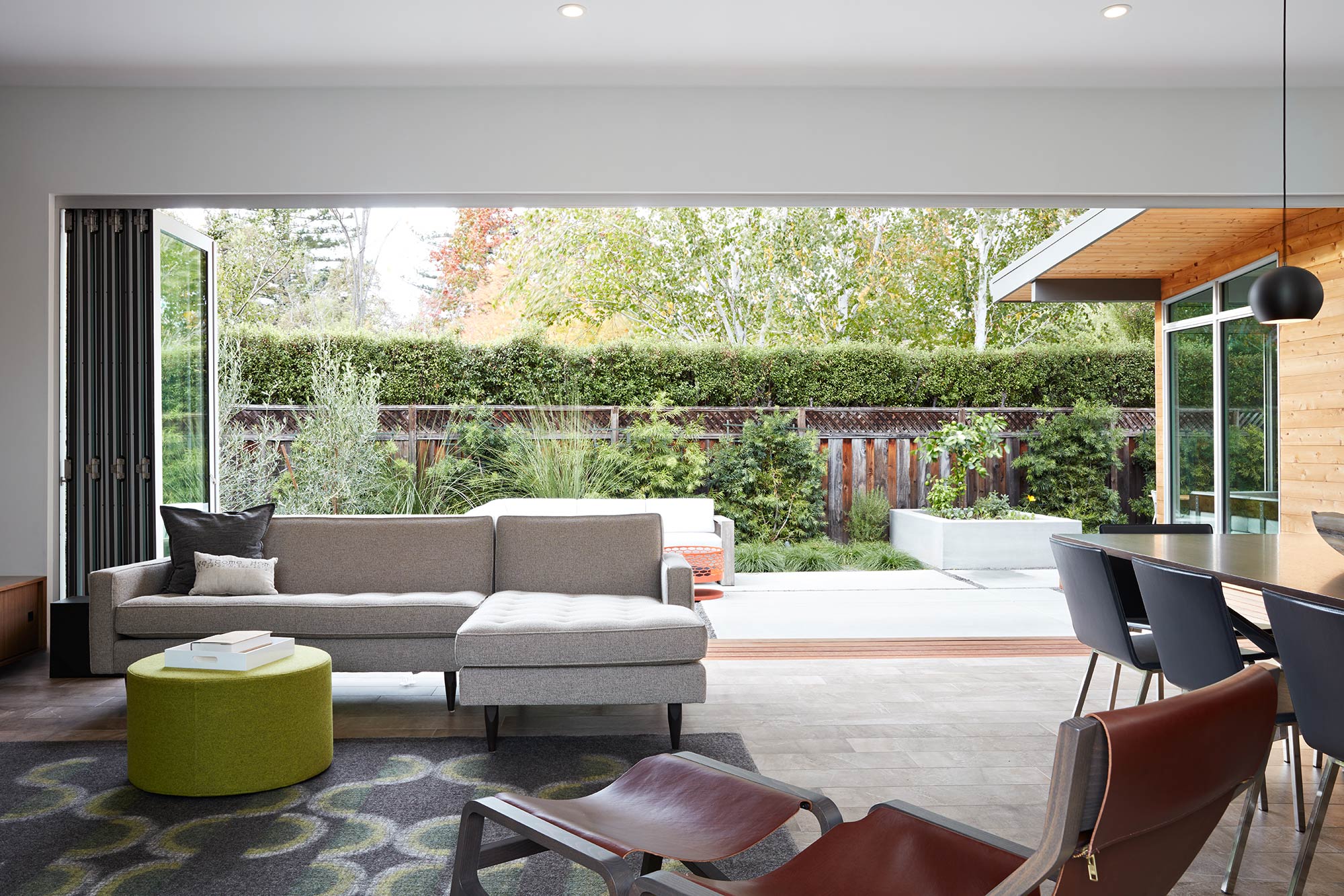 An open, wall-length bi-fold door completely connects the great room to a patio surrounded by lots of plants.