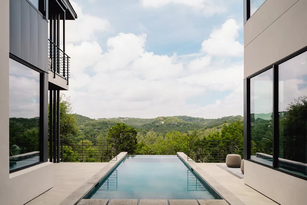 The pool, surrounded by glazing from u-shaped home, overlooks green forestry.