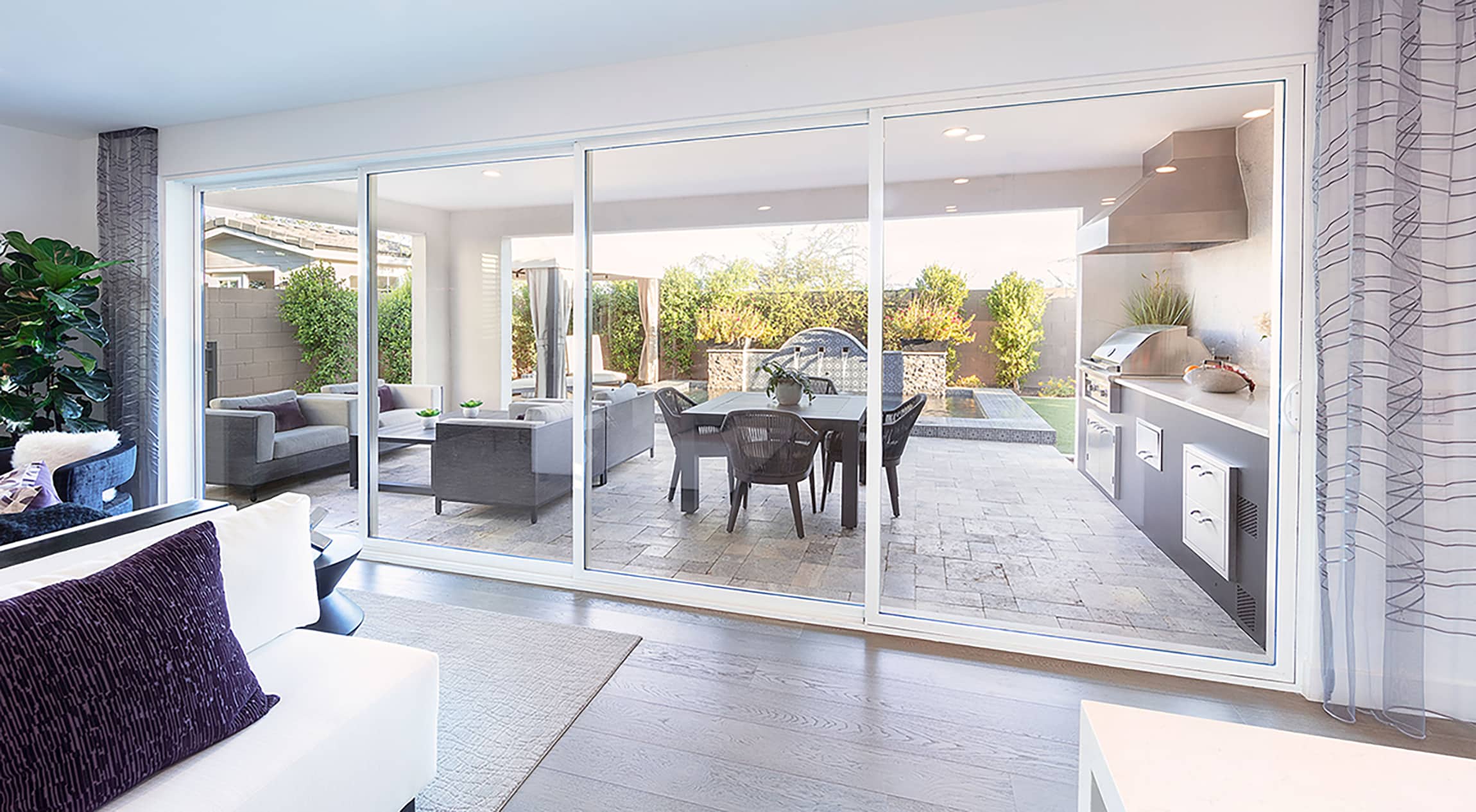 An indoor living room transitions to a furnished outdoor living space through a closed multi-slide glass door.