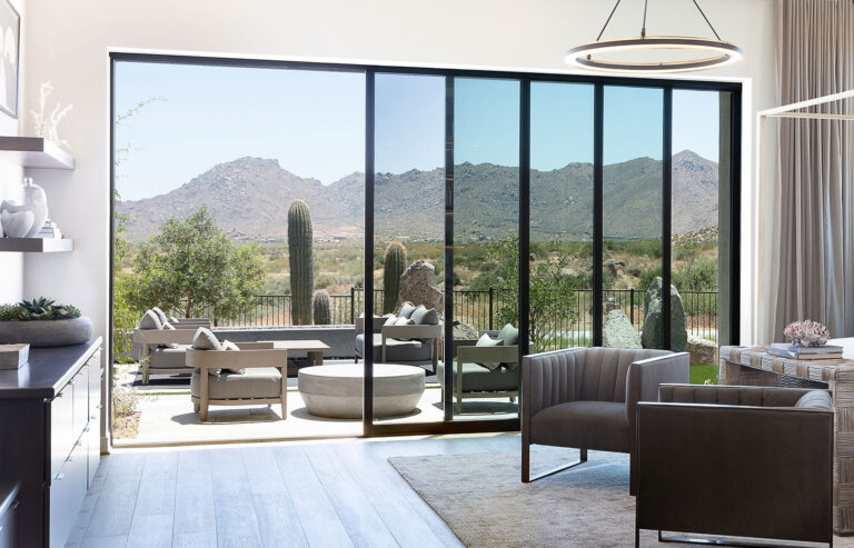 An indoor bedroom and outdoor seating area with a towering mountain view are joined together by a multi-slide glass door.