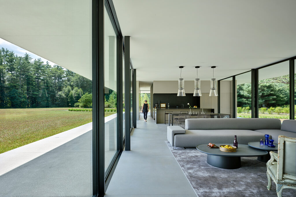 A figure is seen walking down the passage of a glass-walled home. The glass wall makes the surrounding field visible.