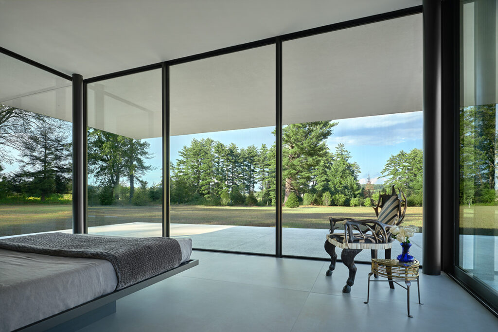 A glass bedroom features huge fixed windows, a modern bed, and concrete floor, contrasted with ornate furniture. Through the window-wall a field is visible.