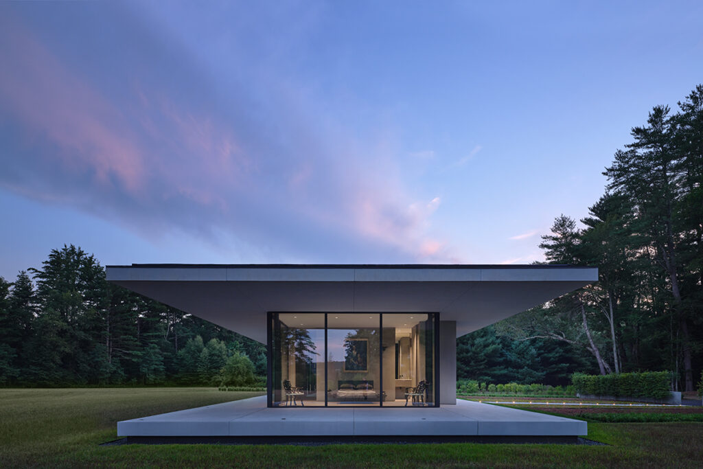 A pavilion-style glass house with an oversized concrete porch and roof overhang sits on a plain at sunset.