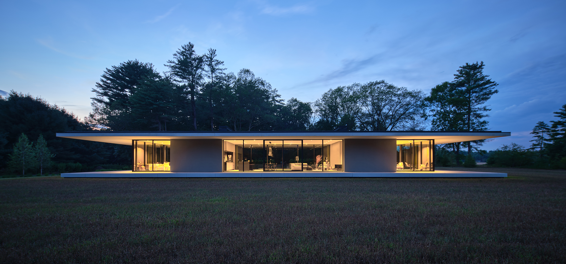 A symmetrical modern glass house sits in a field at dusk; the interior is lit and visible through the large glass windows.