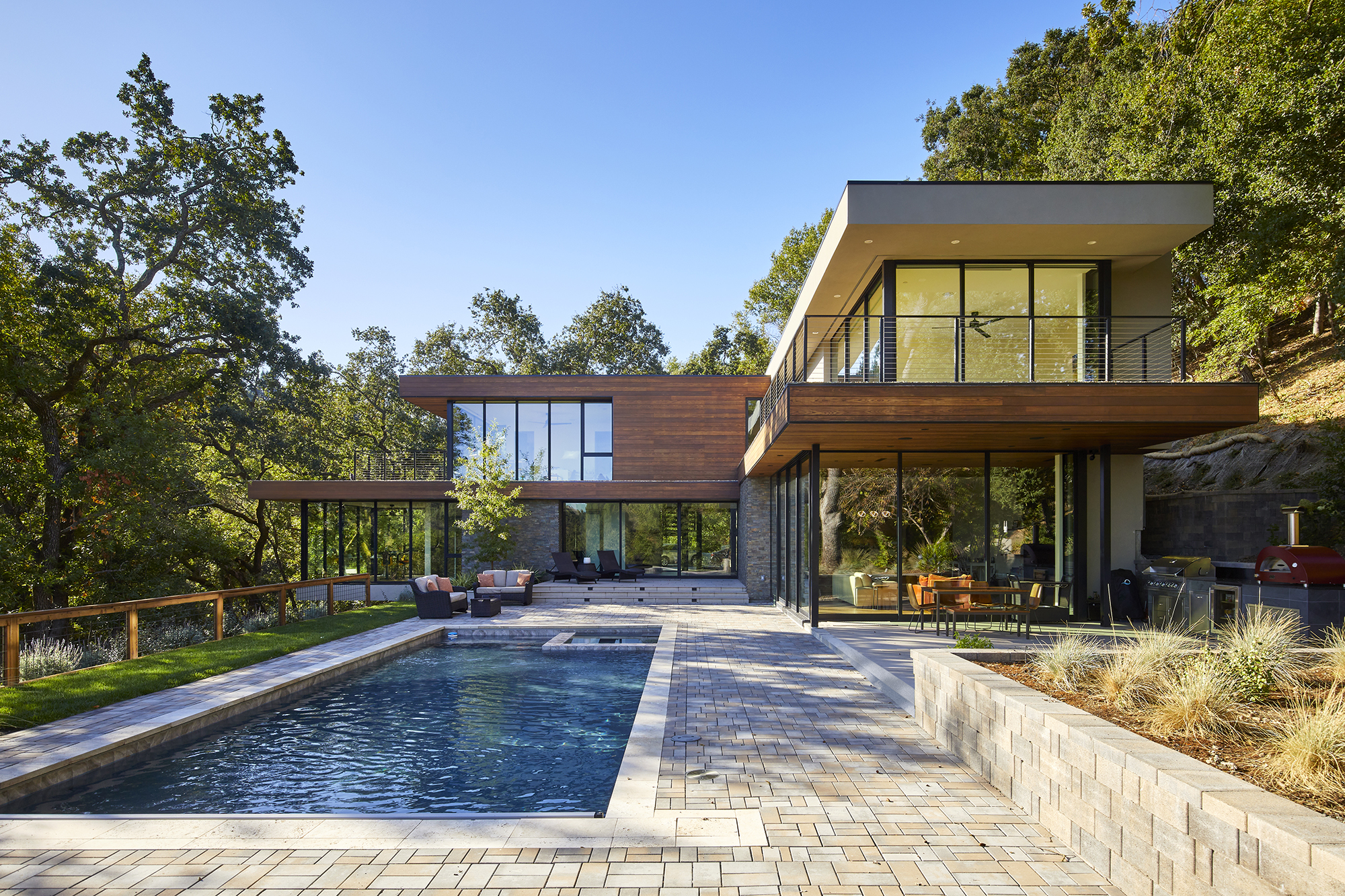 A modern home with floor to ceiling glass walls and horizontal wood slat siding is surrounded by trees. A pool and brick patio with patio furniture is in the foreground.