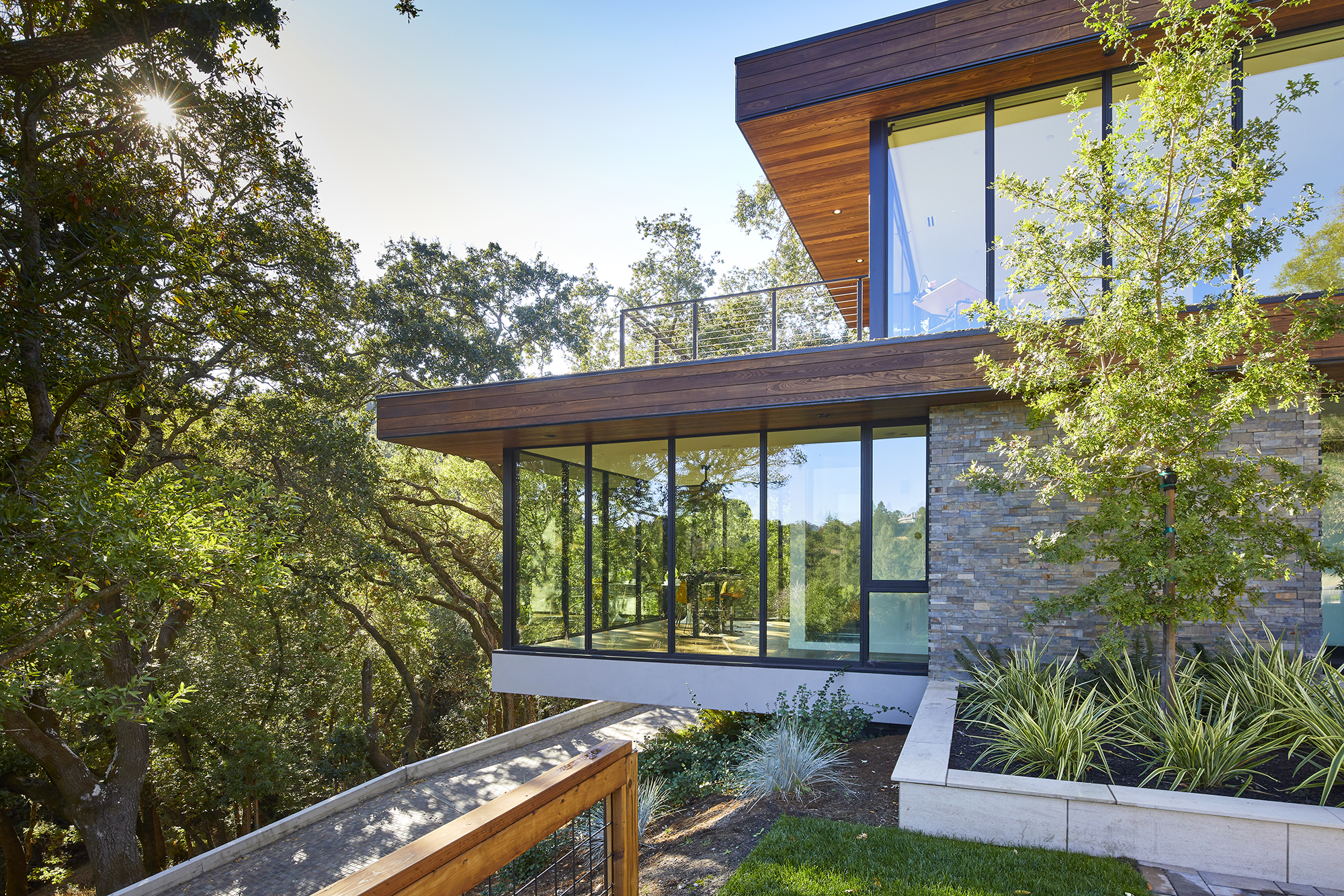 Two stories of a modern home with entirely glass walls reflect the trees across the driveway.