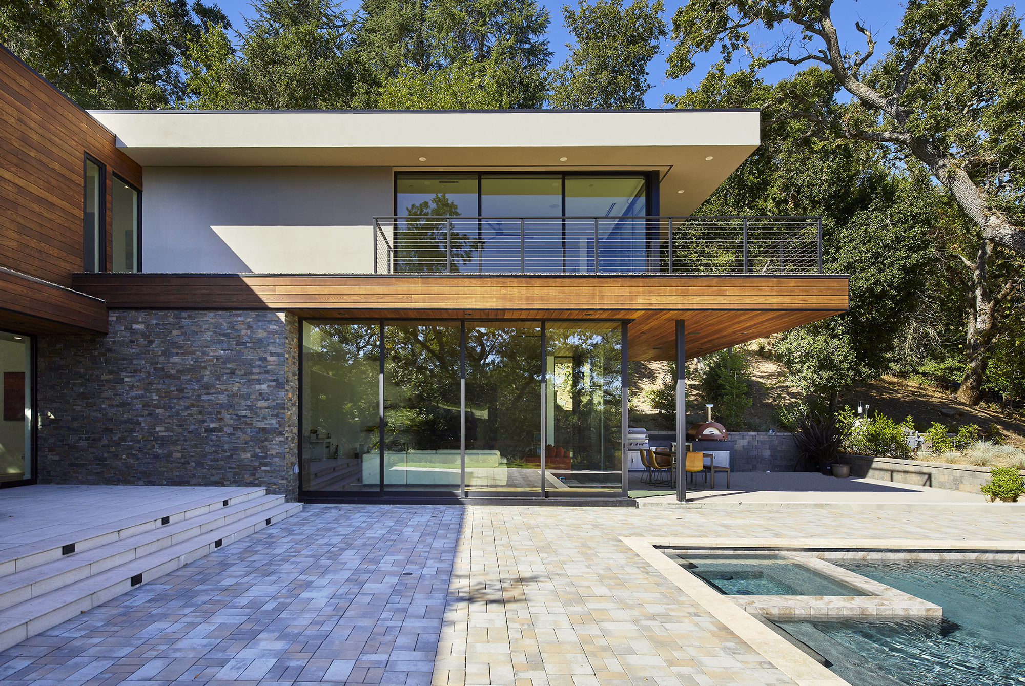 The walls of the first and second story of a modern home are almost entirely glass, and reflect the surrounding trees. A pool and brick patio are in the foreground.