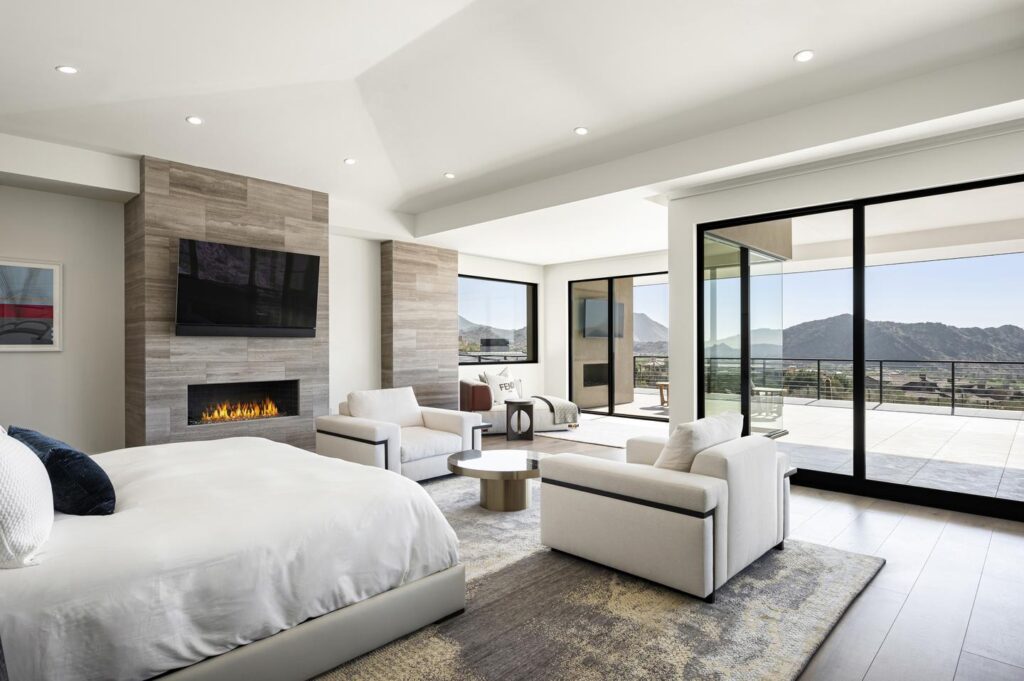 A bedroom is outfitted with a bed, two armchairs, a chaise, a fireplace, a TV, and a glass wall with a sliding door that leads to a balcony. Both the balcony and the glass wall are the length of the room and overlook a neighborhood and desert mountains.