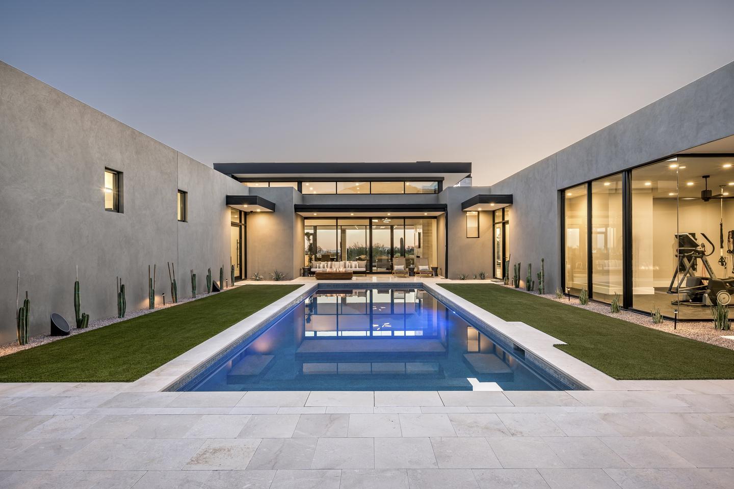 A courtyard with a pool is illuminated by the light coming through the glass walls of the home surrounding it. A portion of the home directly ahead of the viewer is completely transparent due to the glass walls opposite each other, allowing visibility directly through the living room, and out to the sunset on the other side.