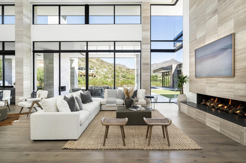 A contemporary living room is set against large glass sliding doors and windows that look out to the backyard and beyond to distant neighbors’ homes, and mountains beyond that.