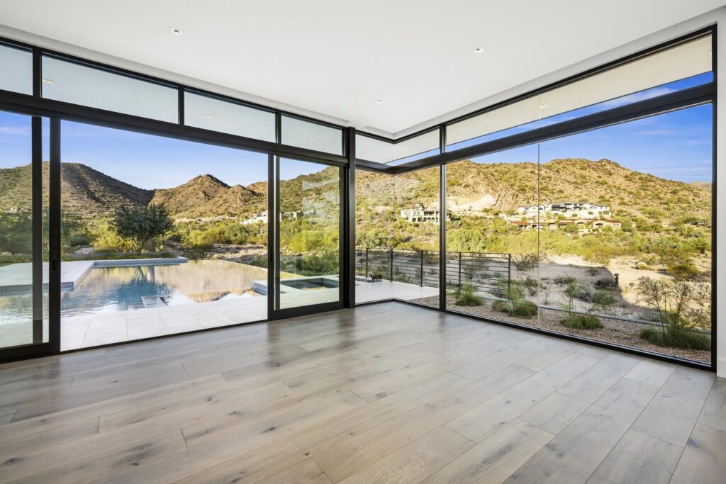 An empty room with wood floors and glass walls overlooks a desert landscape with homes dotting the mountain in the background.