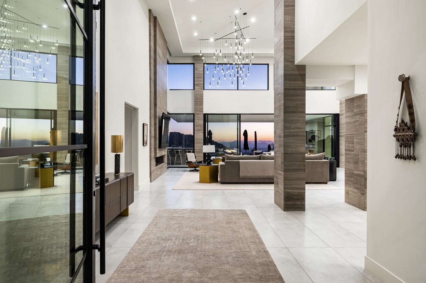 The inside of a home is viewed from the open front door, through a high-ceilinged living room, and out sliding glass doors. The patio furniture is silhouetted against a purple and orange sunset, and purple mountains are in the distance. The high ceiling of the room allows for a dramatic modern chandelier, and large clerestory windows which frame the sky.