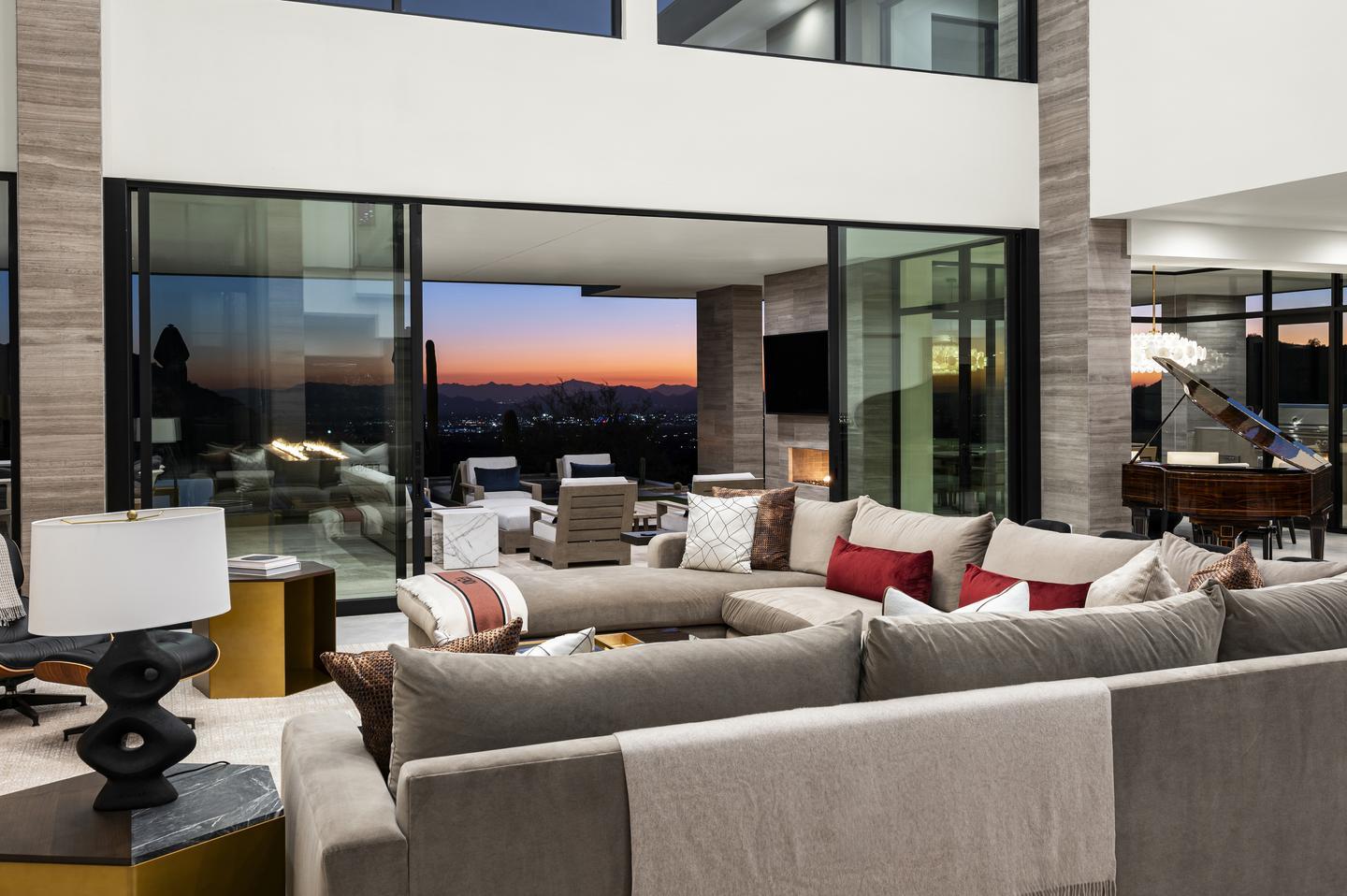 A vivid sunset can be seen through an open bi-parting sliding door, providing a view from the tan sectional in the foreground to the patio, and a cityscape and mountains against the sky.