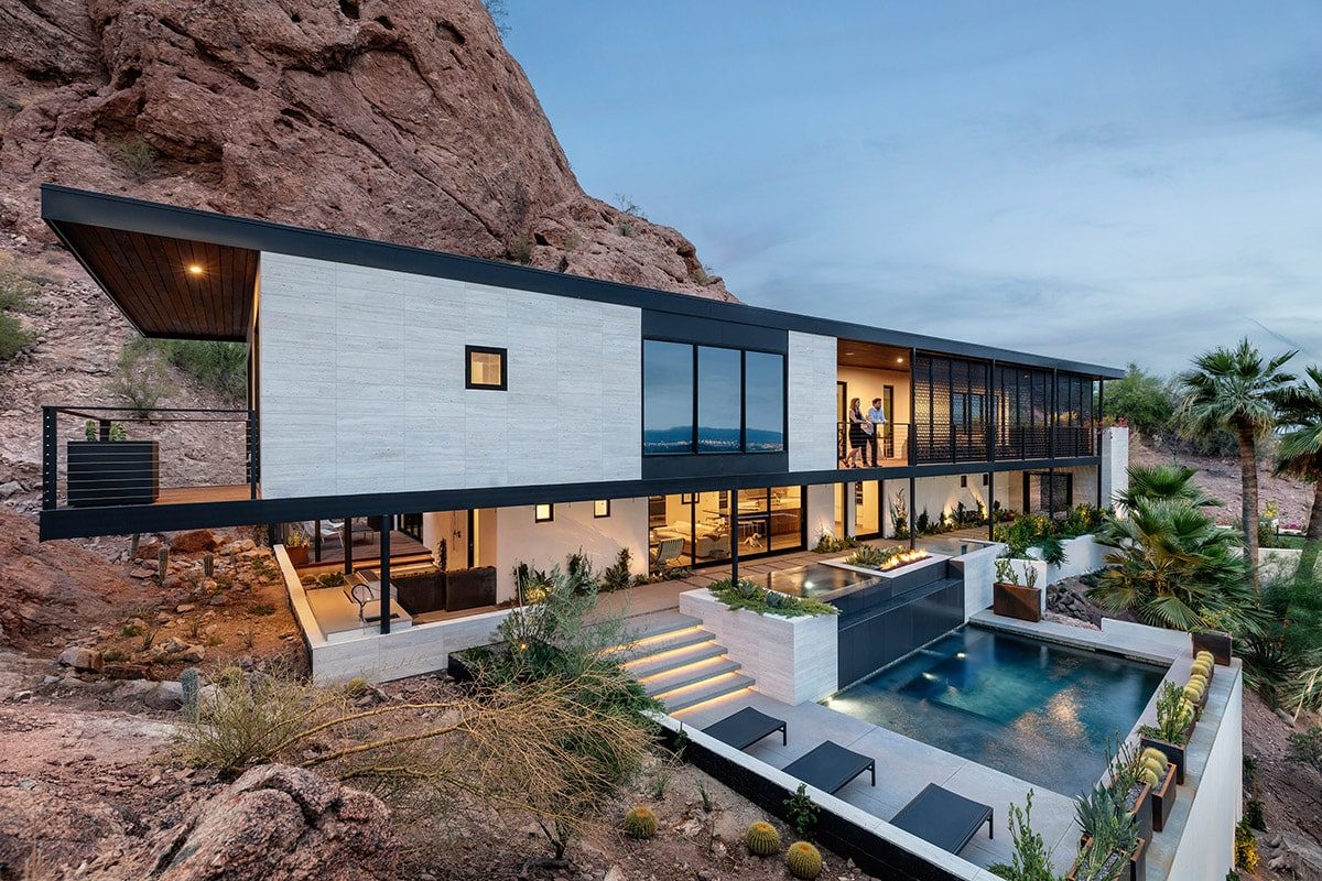 A two-story house built into a mountain side displays a number of floor-to-ceiling window walls and multi-slides looking out onto a pool.
