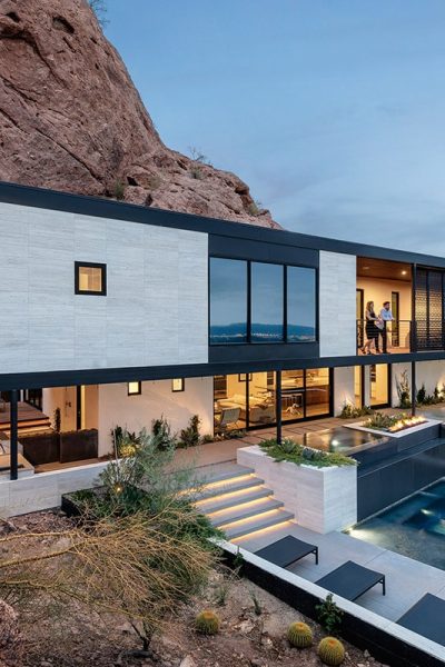 A two-story house built into a mountain side displays a number of floor-to-ceiling window walls and multi-slides looking out onto a pool.