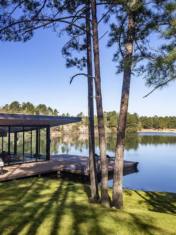 A small glass house sits on a lake, surrounded by trees.