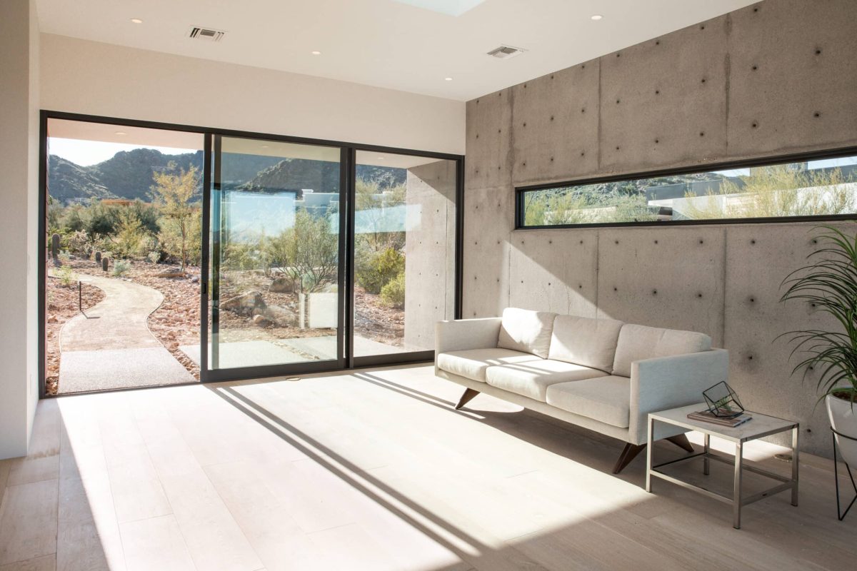 Strong sunlight shining through a glass multi-slide door casts strong shadows on a concrete-walled living room.
