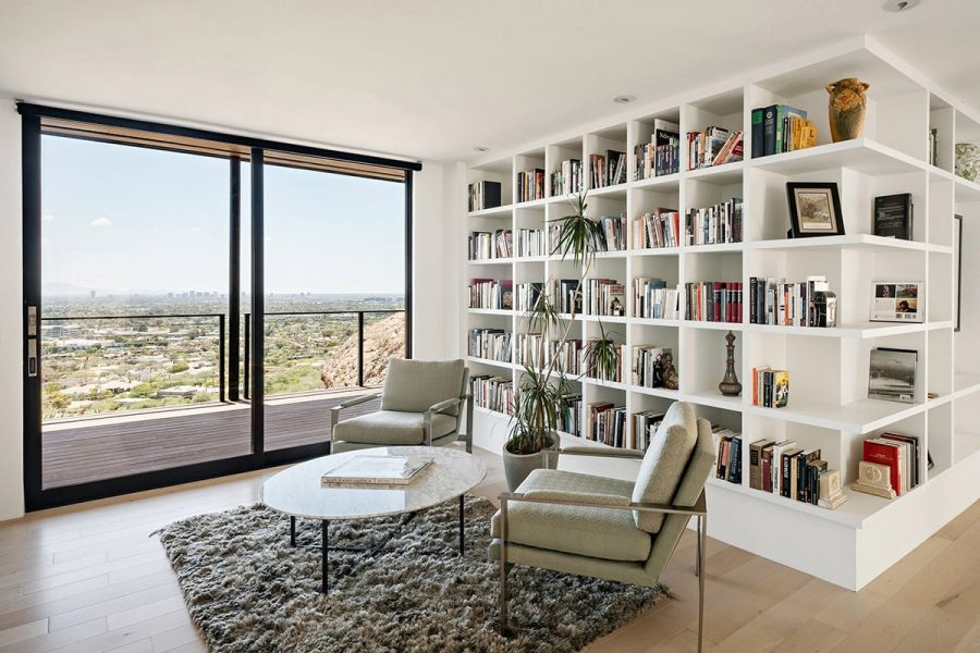 A modern library has white shelves with a tall sliding glass door leading to balcony and view of the desert.