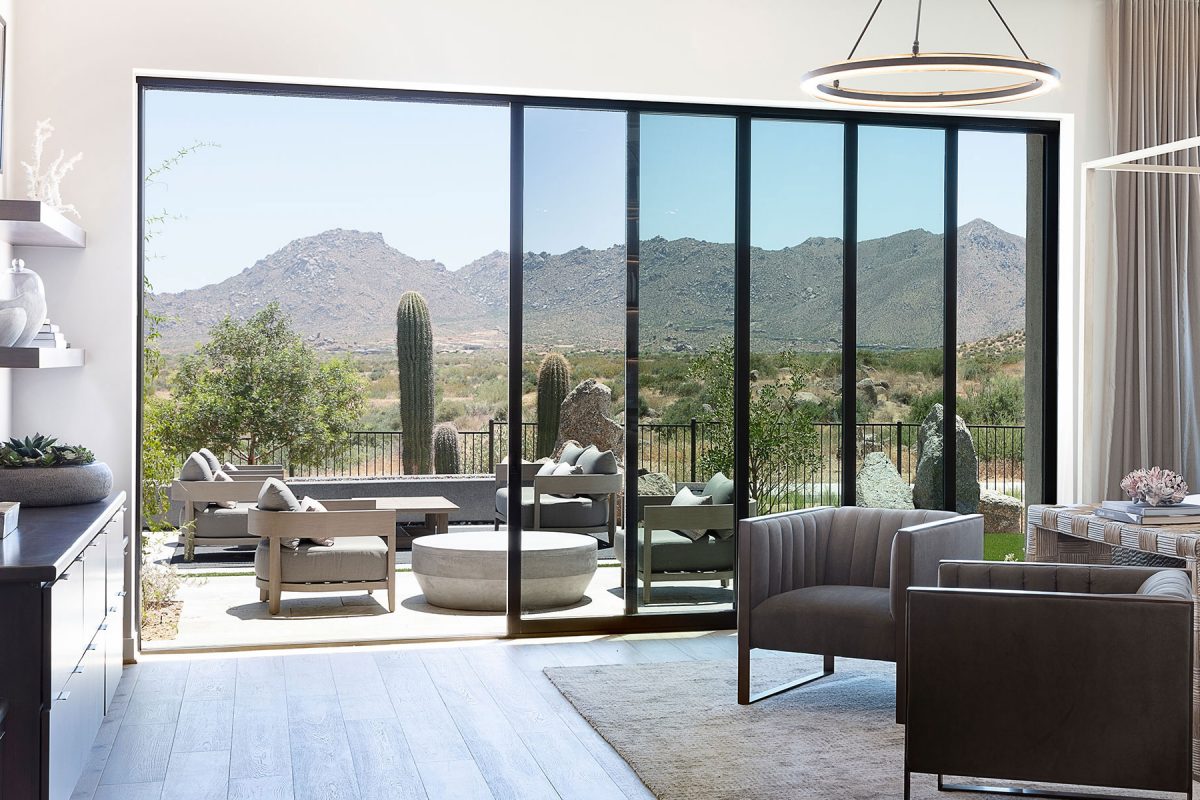 An indoor bedroom and outdoor seating area with a towering mountain view are joined together by a multi-slide glass door.